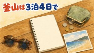 釜山旅行3泊4日の費用のイメージ。木のテーブルにノートと小銭がこぼれた小銭入れ、砂浜と丘を描いた絵はがき、サングラスが置かれている