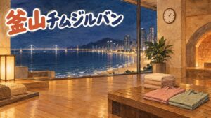 夜の海雲台の浜と光る橋を大きな窓から望むチムジルバンの休憩室を描いた水彩イラスト。木の床に、たたんだタオルとピンクと緑の館内着が置かれ、壁に時計。人物はいない。左上に「釜山チムジルバン」の手書き文字